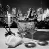 1970s Champagne Bottle In Ice Bucket With Glasses Candle & Napkins On Table & New York City Night Skyline In Background view 1